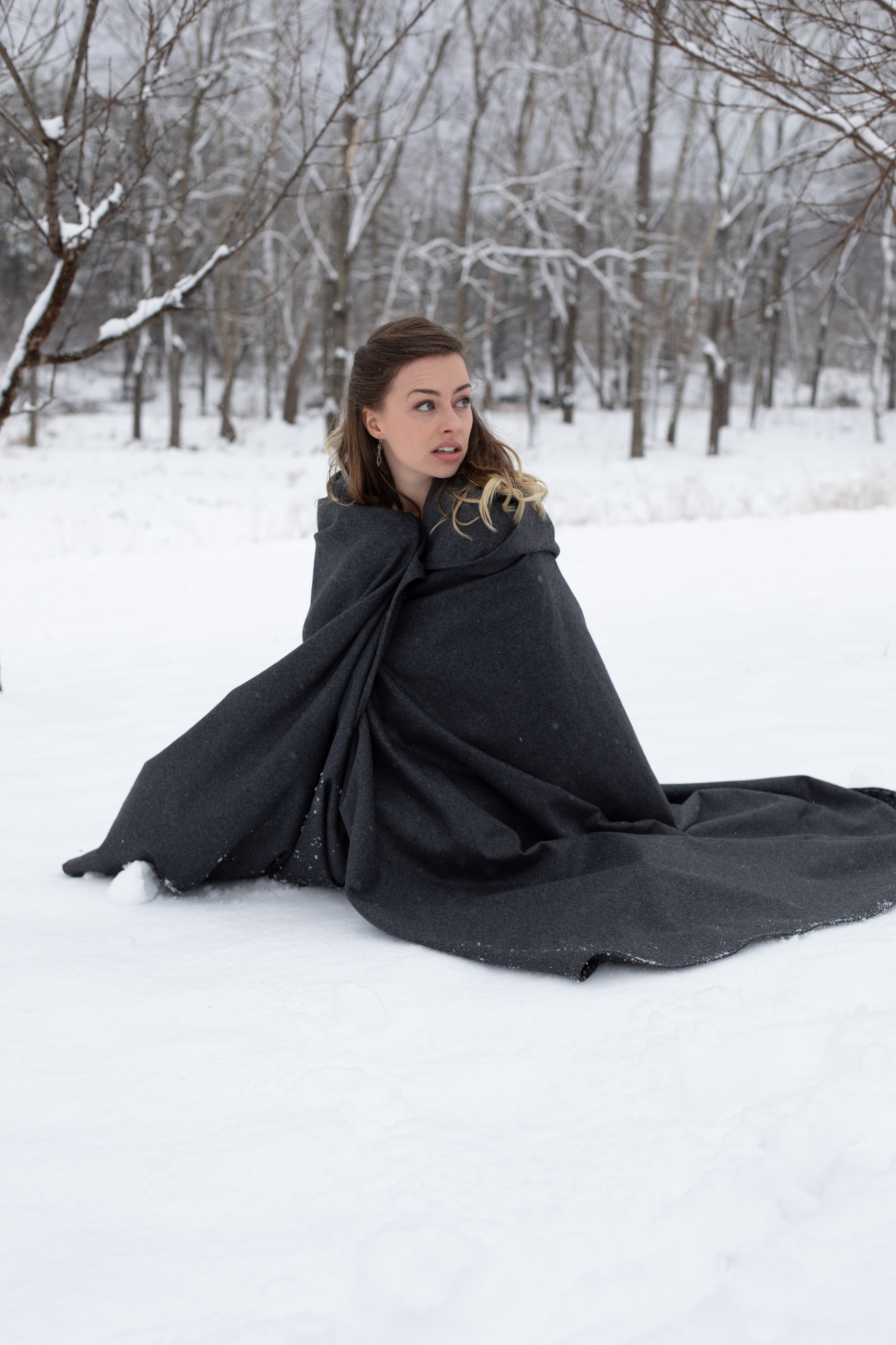 a young woman with long brown hair is huddled on the ground in the snow, wrapped in a dark gray blanket. she is looking off to the right side of the frame with a surprised or concerned expression. the background is a snowy forest with bare trees.