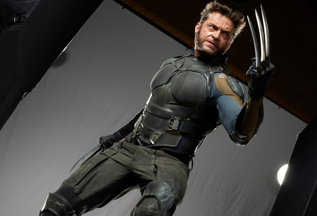 a lowangle shot of hugh jackman as wolverine in a dark gray and black superhero suit, with his signature metal claws extended, standing in an action pose on what appears to be a movie set.