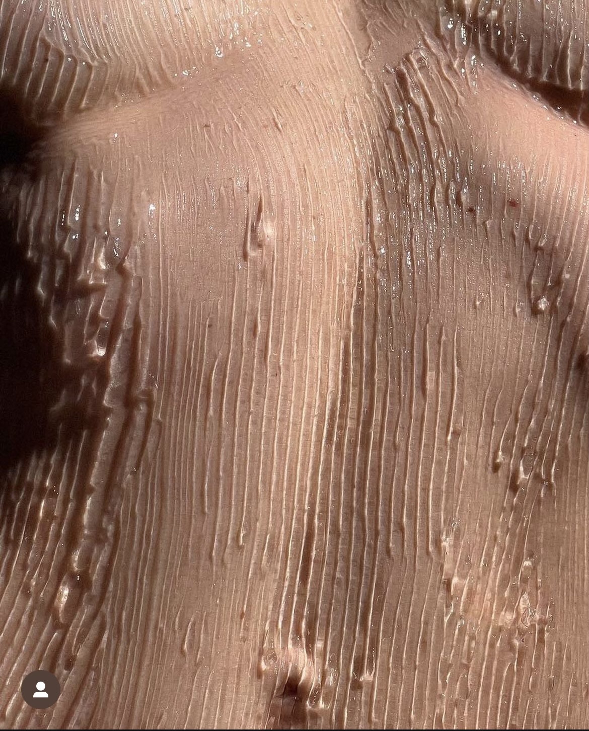the image is a closeup, macro shot of a person's torso.  the skin is covered in a thick layer of a light beige, glossy cosmetic product, possibly a cream, lotion, or serum. the product has been applied in vertical streaks creating a textured, ridged pattern across the skin. the lighting enhances the glossy sheen of the product, highlighting its texture and consistency. the overall appearance is smooth and hydrated. the focus is entirely on the skin and the product's texture. the image has a somewhat artistic or aesthetic quality.