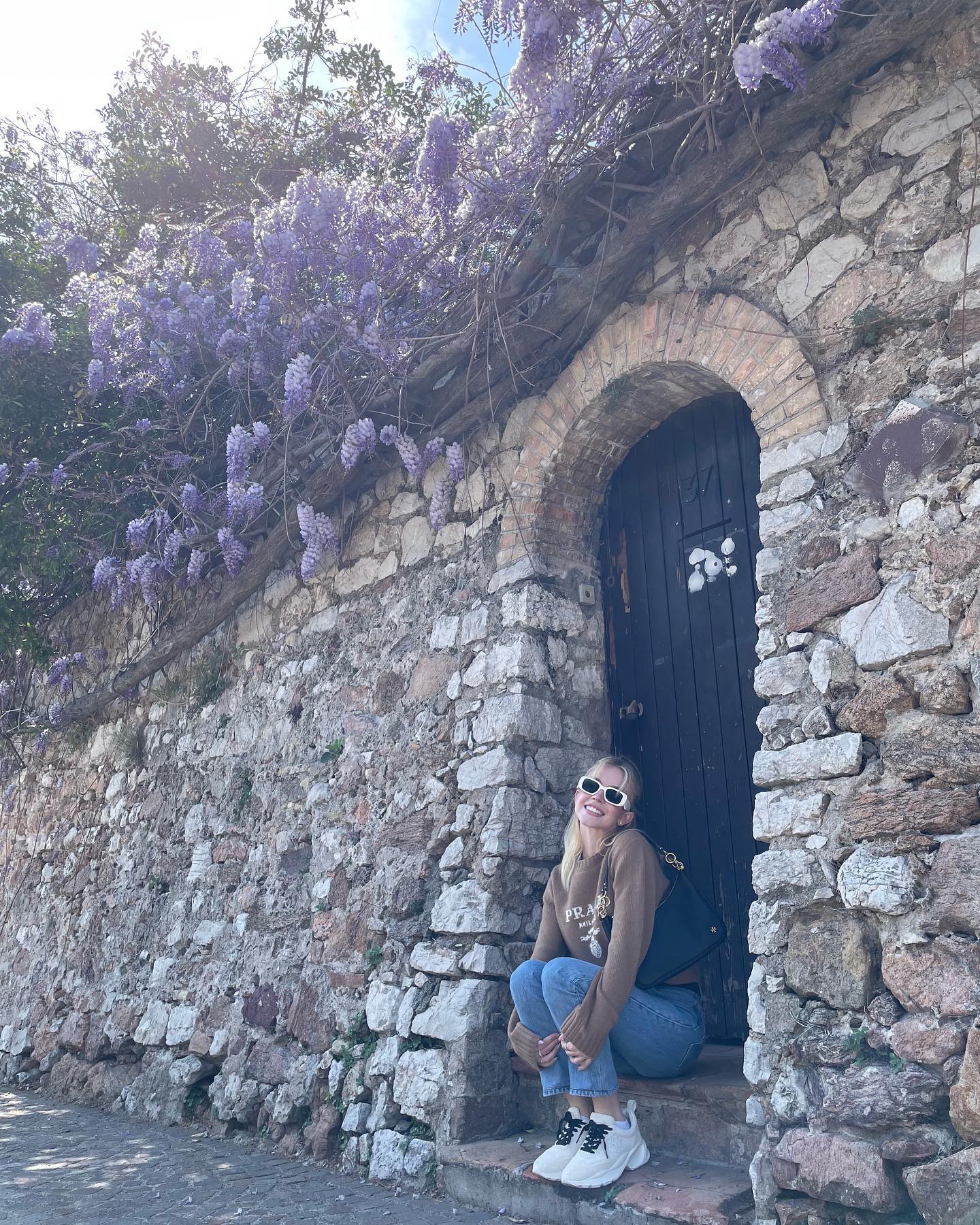 a young woman with sunglasses sits smiling on the steps of an old stone doorway, which is covered by a vibrant wisteria vine in full bloom. she is wearing a brown sweater, jeans, and white sneakers. the scene is outdoors, sunny and picturesque.  the stone wall is made of irregular, aged stones. the overall feel is one of spring, travel, and happy relaxation.