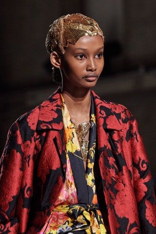loseup shot of a fashion model showcasing a red floralpatterned coat and a gold headpiece. she is wearing a vibrant floral kimono underneath and gold necklaces and earrings. the background is dark and blurry, focusing attention on the model and her attire. the image appears to be from a fashion show or photoshoot.