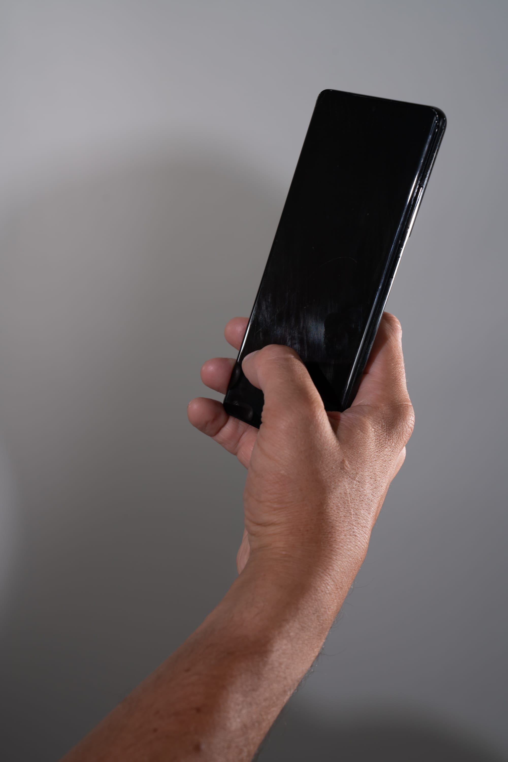 a man holds a smartphone in front of him against a plain grey background. the phone is black and the screen is blank. the man's arm is extended and his hand is gripping the phone. the lighting is artificial, creating a soft shadow behind the man's arm.