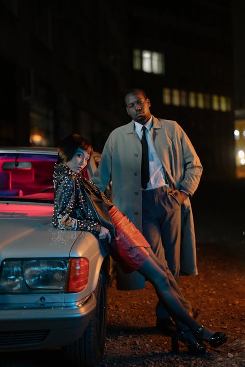 a stylish woman in a studded leather jacket and red mini skirt leans against a vintage car. a man in a trench coat stands beside her, creating a mysterious and fashionable scene at night.  the urban backdrop and lighting contribute to a film noir aesthetic. the couple's poses and attire suggest a sense of confidence and perhaps a hint of intrigue.