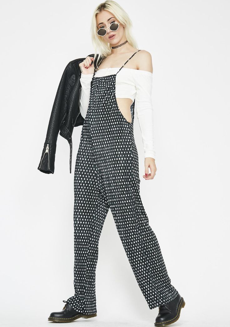 a woman stands in front of a white background. she is wearing sunglasses, a black choker necklace, a white offtheshoulder longsleeved top, and black patterned overalls. she is also wearing black boots. she is holding a black jacket on her left shoulder.