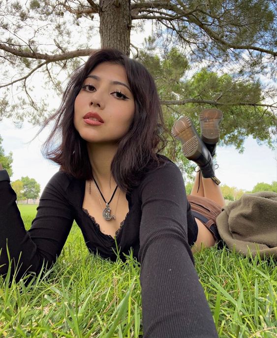 a young woman with dark hair and makeup is taking a selfie while laying in the grass under a tree. she is wearing a black long sleeve shirt, brown corduroy pants, and black doc martens. she is also wearing a necklace with a crescent moon pendant. the photo appears to be taken in a park on a sunny day. the lighting is natural and soft. the perspective is from the woman's point of view, as it is a selfie.