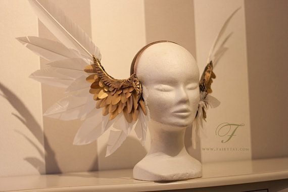 a closeup shot of a white mannequin head adorned with a unique handmade headband, showcasing the delicate detail of intricate golden wings made of feathers.  the wings are crafted with both real white and gold feathers, adding a touch of elegance and whimsy to the design.  the headband itself appears to be a simple white band, allowing the focal point to be the intricate wings. the background is a simple white and brown striped wall. the mannequin head is positioned on a white shelf, creating a clean and minimalist aesthetic. the overall effect is ethereal and enchanting.