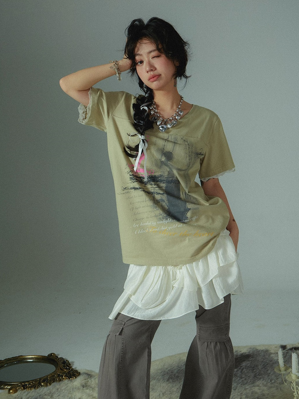 a young woman poses for a fashion photograph. she is wearing a light beige graphic tshirt with lace detailing at the sleeves. the tshirt features an artistic print.  she's also wearing a layered white ruffled skirt over gray wideleg pants, creating a layered, unique look. she has on silver jewelry, a necklace and a bracelet, and her hair is styled in a braid. her expression is playful, winking at the camera. the background is a simple, lightcolored studio backdrop.  she appears trendy and confident.