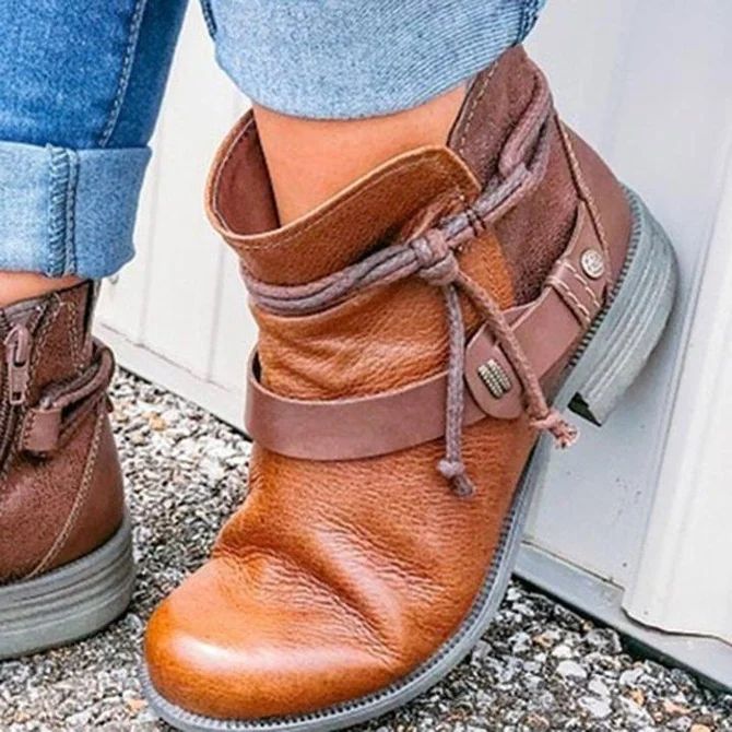 a close up of a woman wearing brown leather ankle boots with a decorative lace. the woman's feet are angled out, so only the right boot is visible.  the boots have a rounded toe and a chunky heel. they are made of a brown leather and have a brown strap that goes around the ankle. the strap is decorated with a metal buckle. the boots are being worn with a pair of blue jeans. the background is blurred.