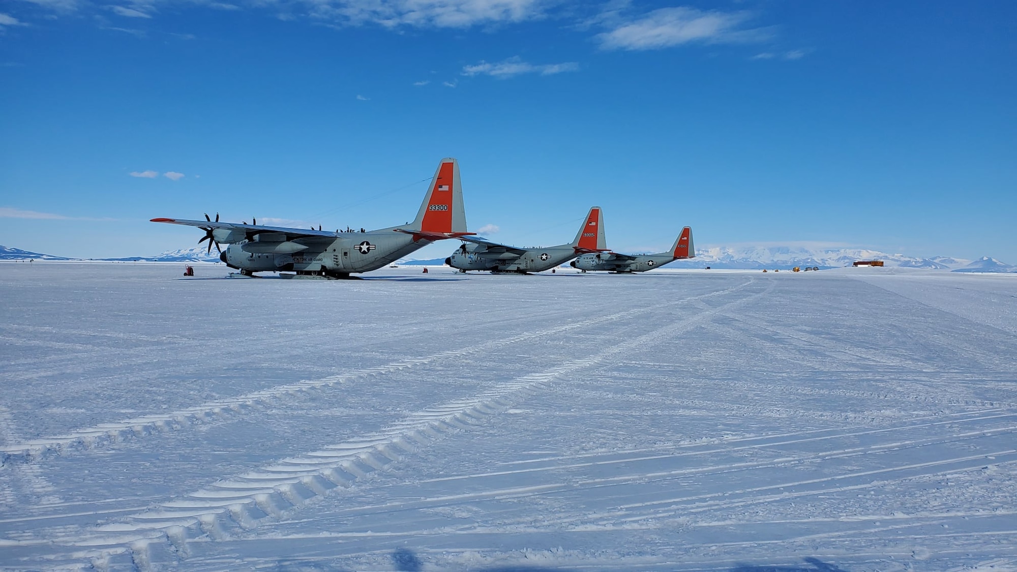 three gray military cargo planes are parked on a snowy runway in antarctica. the sky is blue with clouds.  a small, red building is on the right side of the image.