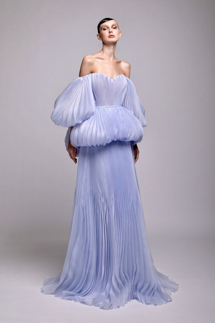 a woman in a light purple, offtheshoulder pleated gown with large, puffy sleeves and a flowing skirt. the dress is formal and elegant, and the woman is posed against a neutral background.