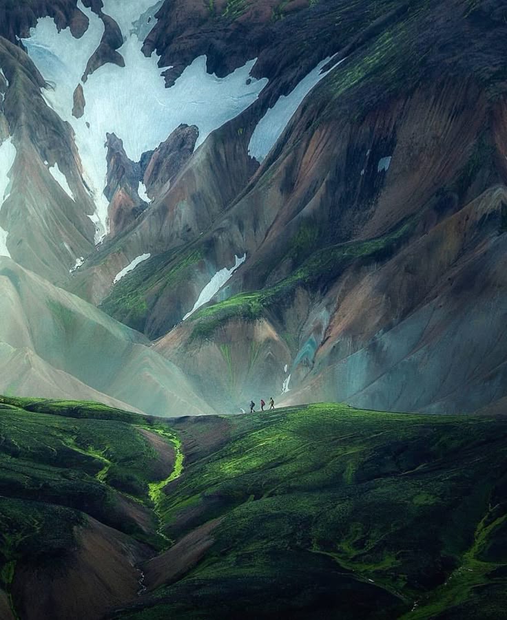 the image captures a vast, mountainous landscape, likely in iceland. towering, colorful mountains with patches of snow reach towards the sky. in the foreground, a group of three hikers can be seen walking along a green hill. a small stream of bright green vegetation cuts through the hillside leading towards them. the scene conveys a sense of adventure, isolation, and the beauty of the wilderness.