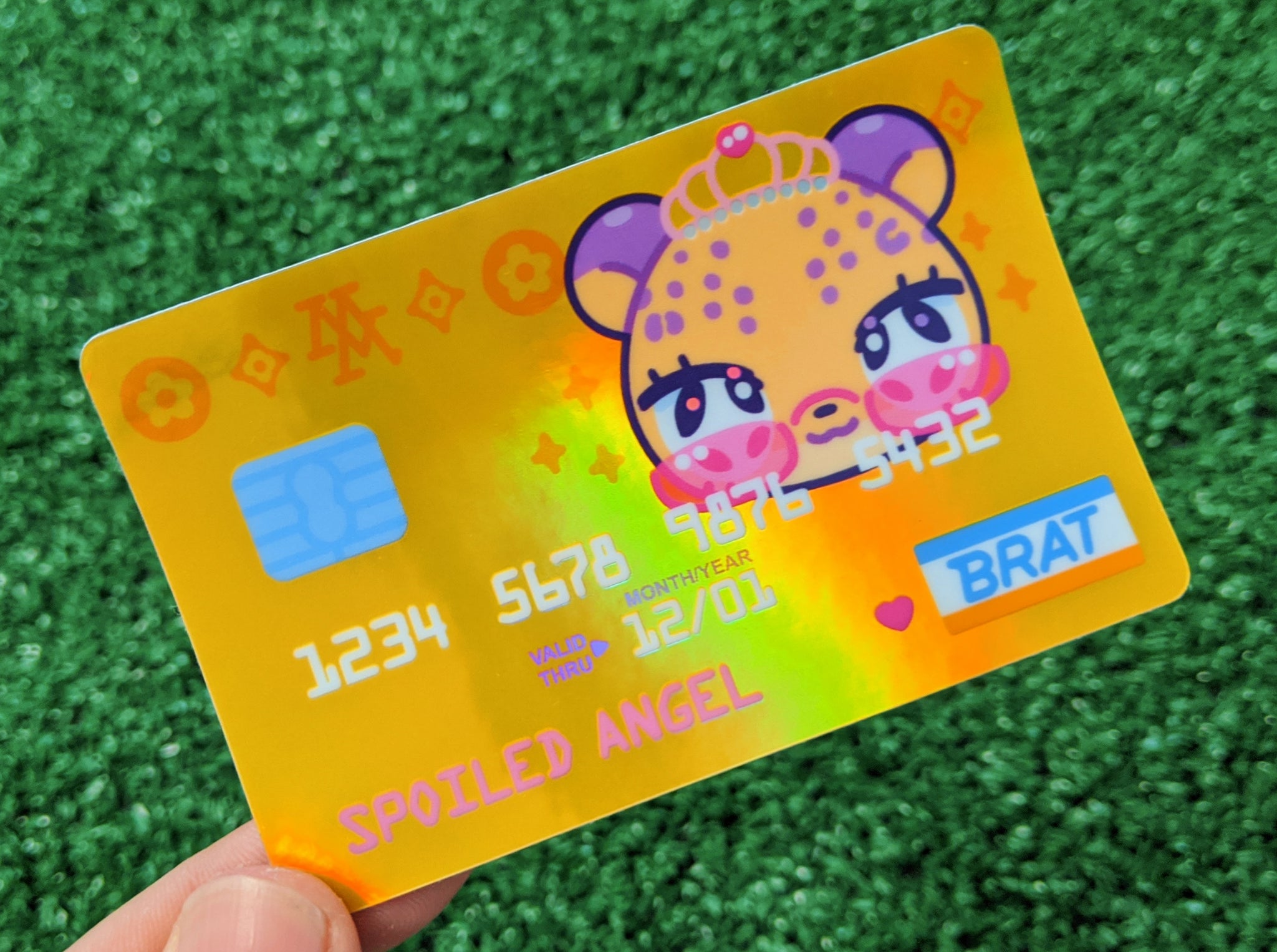 closeup shot of a holographic sticker designed to resemble a credit card. the card features a cute cartoon animal character with a crown, surrounded by various decorative elements. the design is vibrant, with a mix of yellow, pink, and purple tones. the text "spoiled angel" and "brat" are visible, along with simulated credit card numbers and expiry date. the sticker is held in a hand against a green background.  the overall style is playful and reminiscent of kawaii or y2k aesthetics.