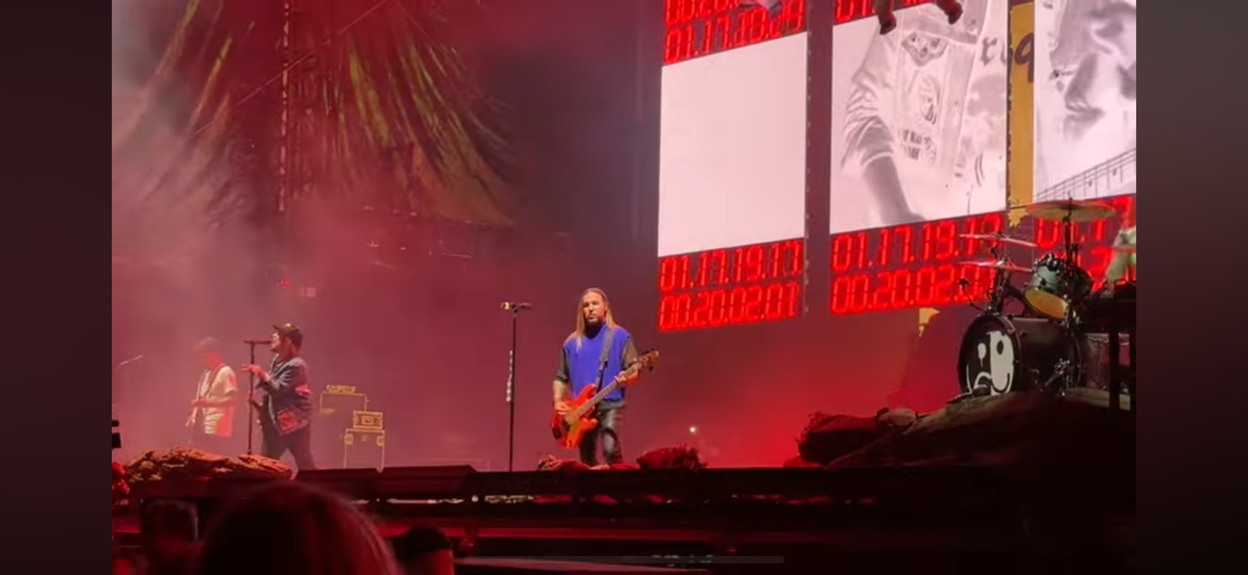 a rock band is performing on stage in front of a large crowd. the band consists of a guitarist, a drummer, and a singer. the stage is lit with red lights and the band is playing their instruments. the crowd is cheering and enjoying the performance. the guitarist is wearing a blue shirt and the drummer is wearing a black shirt. the singer is wearing a white shirt and a black jacket. the band's equipment is set up on the stage, including microphones, drums, amplifiers, and speakers.