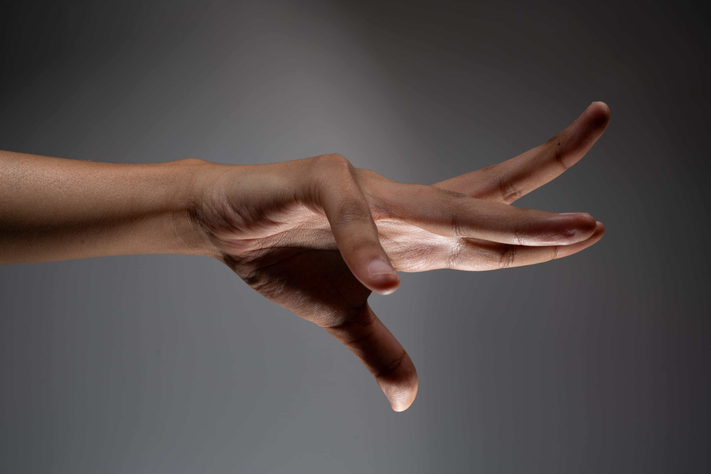 the image shows a hand with a dark complexion against a dark gray background. the hand is positioned with the palm facing the camera and the fingers extended and slightly separated, creating a dynamic and expressive pose. the lighting is dramatic, casting shadows that highlight the contours of the hand.