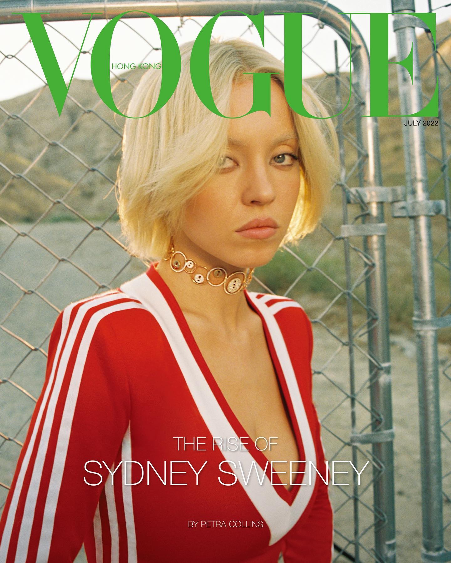 the image shows the july 2022 cover of vogue hong kong featuring actress sydney sweeney.  sweeney is the central focus, posing in a red adidasstyle tracksuit with white stripes. she has blonde, shoulderlength hair and is wearing a gold and button choker necklace. the background is a wire fence, suggesting an outdoor setting. the vogue logo is prominently displayed in green,  and the coverline reads "the rise of sydney sweeney," crediting petra collins as the photographer. the overall aesthetic is retro and slightly vintage.