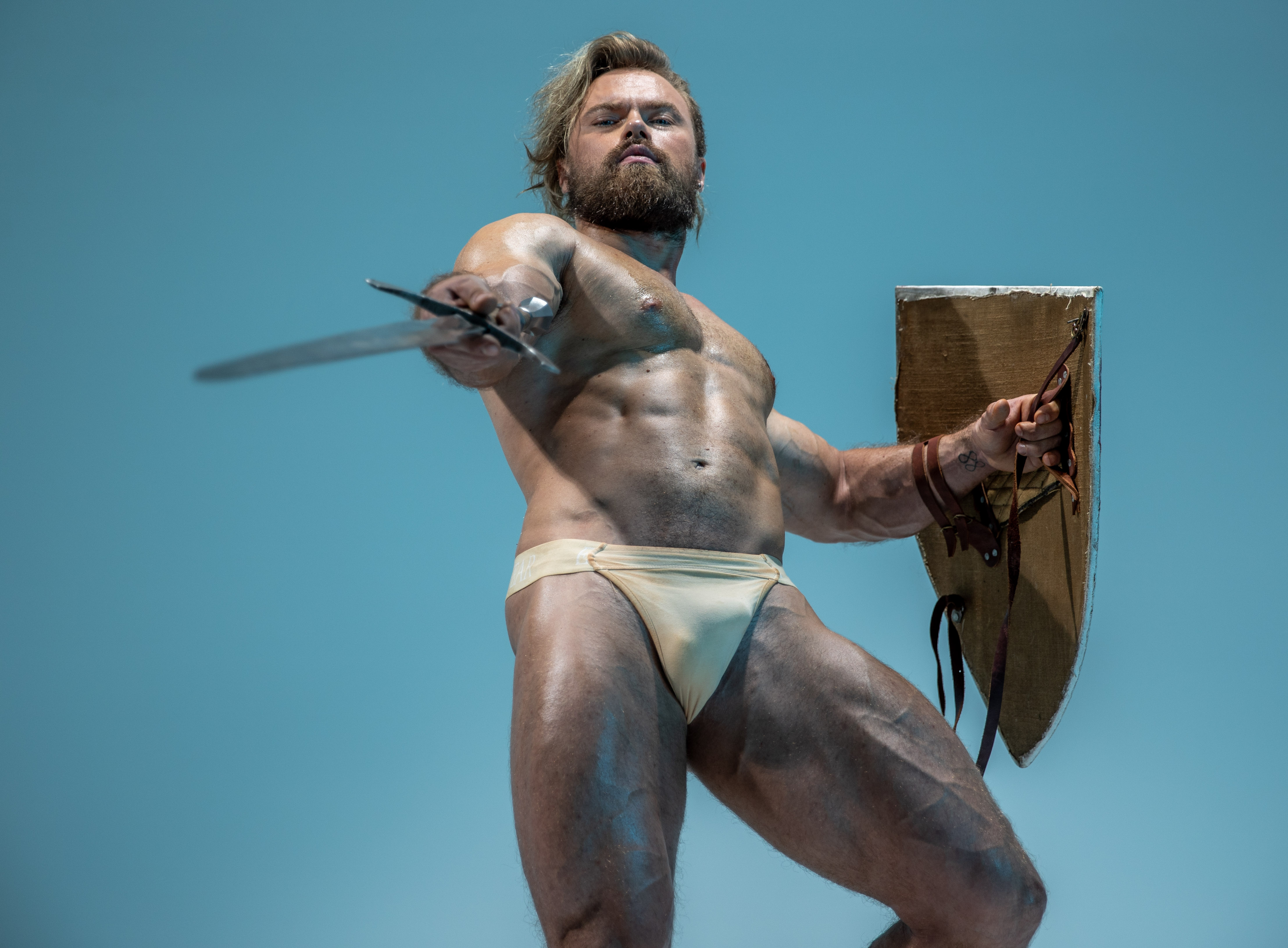 a muscular, bearded man stands in a powerful pose against a light blue background. he holds a sword outstretched in his right hand and a large, brown shield in his left. his body glistens, suggesting oil or sweat, further emphasizing his muscular physique. the low angle shot adds to the perception of power and dominance.