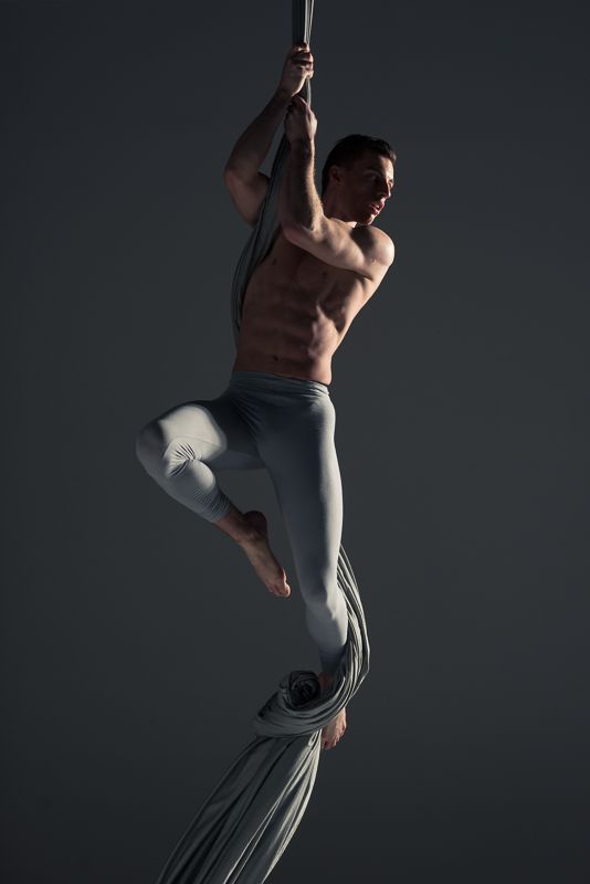 a man is performing an aerial silks routine, hanging from a gray fabric suspended from the ceiling.  he's shown with his legs outstretched and his torso contorted in a challenging pose. he appears to be midroutine, with his legs extended and his arms engaged in holding the silks as he appears to be balancing midair.  the photograph was likely taken in a studio,  under low lighting.  the background is a dark gray, and the lighting casts dramatic shadows on his body. the man is clad in grey workout pants, showing off his athletic physique and the strength and flexibility required for this challenging art form.