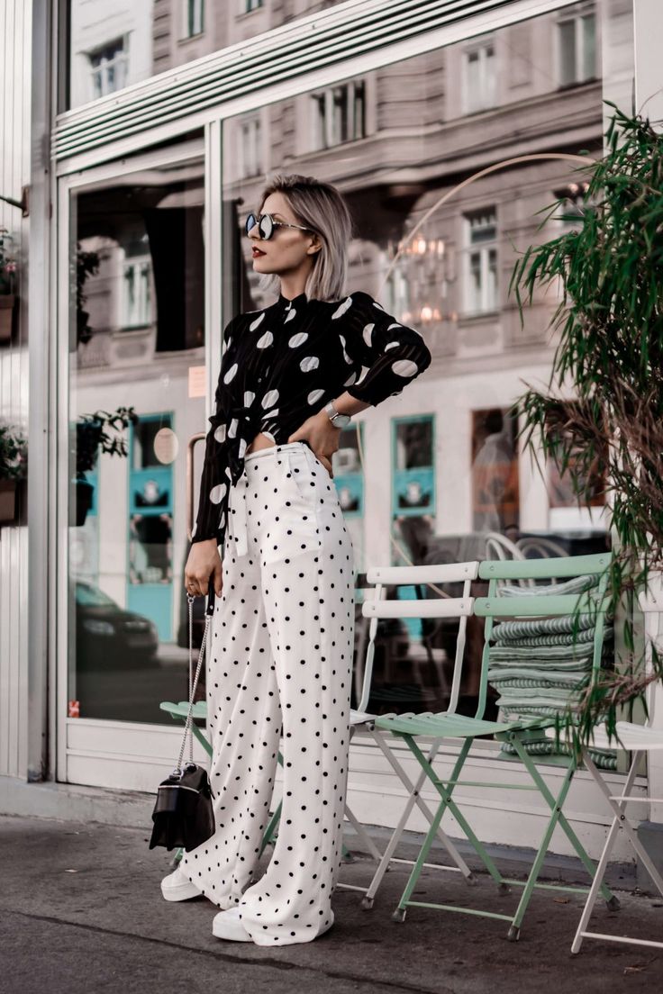 a stylish woman is standing outdoors in front of a cafe. she is wearing a black and white polka dot blouse and matching wideleg palazzo pants.  she also has on sunglasses and a black handbag.  the setting appears to be a city street with a cafe in the background. the overall style is chic and elegant, showcasing a fashionable summer outfit.