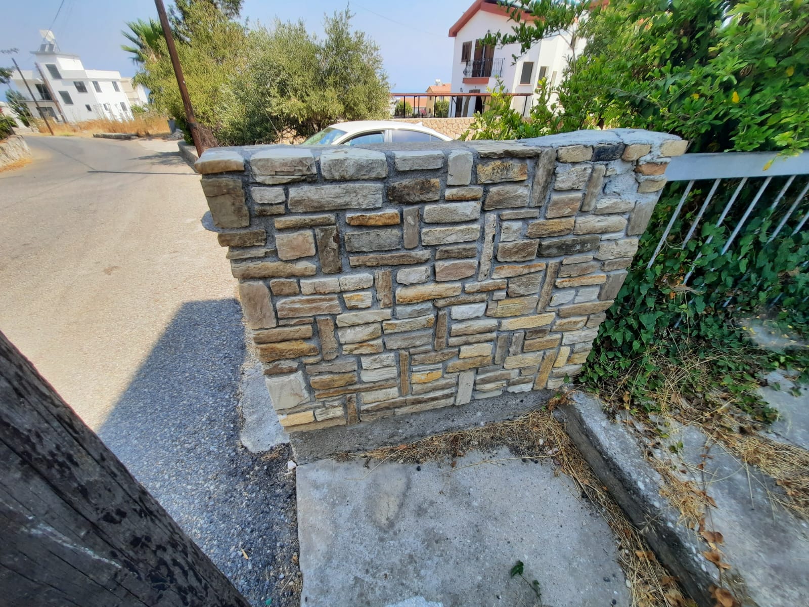 the image shows a stone wall next to a road and a curb. there are plants and bushes behind the wall, and buildings and a car can be seen in the background. a pole is on the left side of the image.