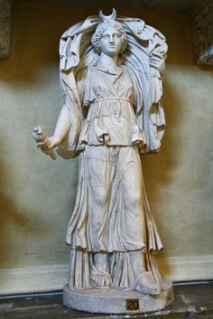 a fulllength marble statue of a woman, likely a roman goddess, standing on a pedestal. she is wearing a flowing robe and a crescent moon adorns her head. her right hand holds a torch and her left hand rests on a draped cloth. the statue is welllit, showcasing the intricate details of the drapery and the woman's serene expression.  the background is a simple wall with a yellowish hue, drawing focus to the white marble sculpture.