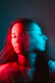 a woman's face is partially obscured by a blur of red and blue light. the background is dark and the colors create a mysterious and ethereal atmosphere. the image appears to be a double exposure with the woman's face visible in the center but blurred and distorted by the bright lights. the overall effect is one of mystery and intrigue.