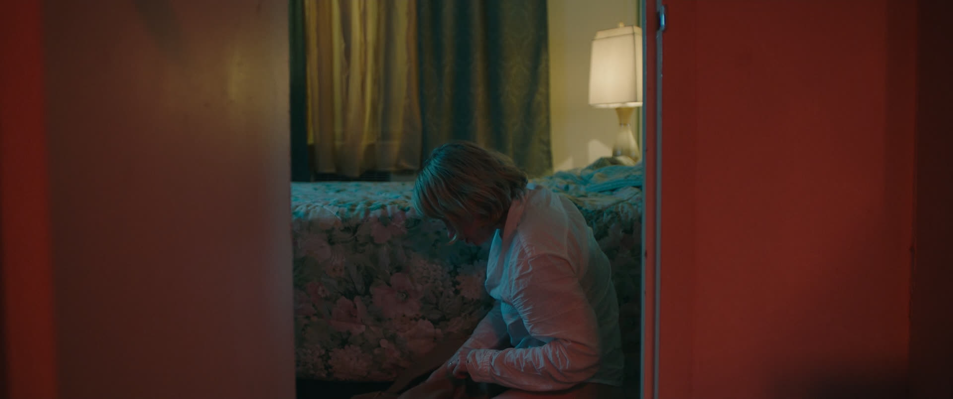 a woman sits on the floor of a hotel room, illuminated by a single lamp. the room is dimly lit and has floral patterned bedding. the woman is dressed in white, and appears to be alone. she is staring at the floor, and looks sad and lonely.