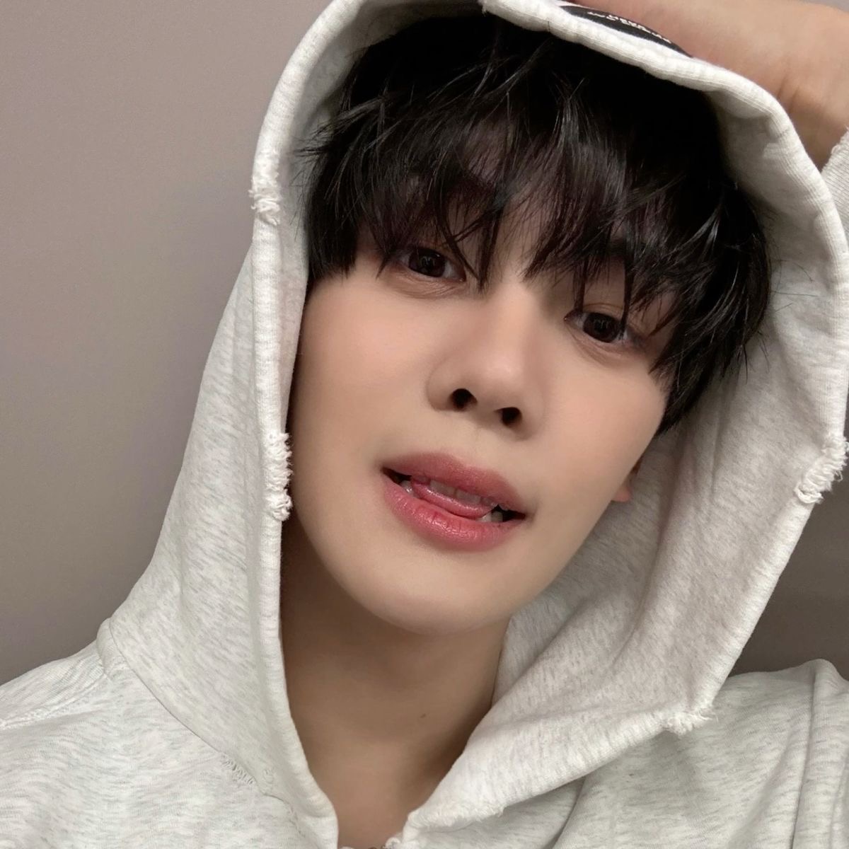 the image is a closeup selfie of a young asian man. he is wearing a light grey hoodie and has his hand resting on the hood. he has dark hair styled with a fringe, and his lips are slightly parted in a subtle expression. the lighting is soft, creating a natural and flattering look. the overall feel of the image is casual and intimate.