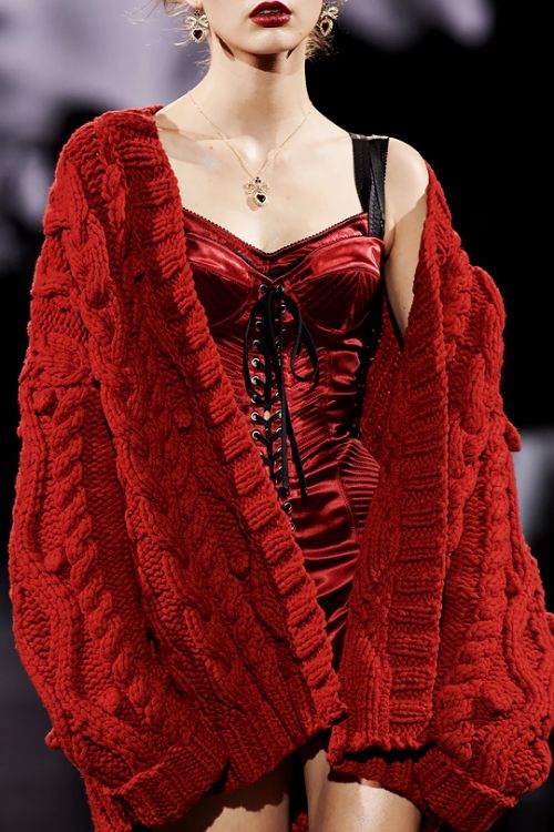 a fashionable model is wearing a red cable knit cardigan over a red satin corset dress, she is also wearing gold jewelry, earrings and a necklace, her lips are a bright red.