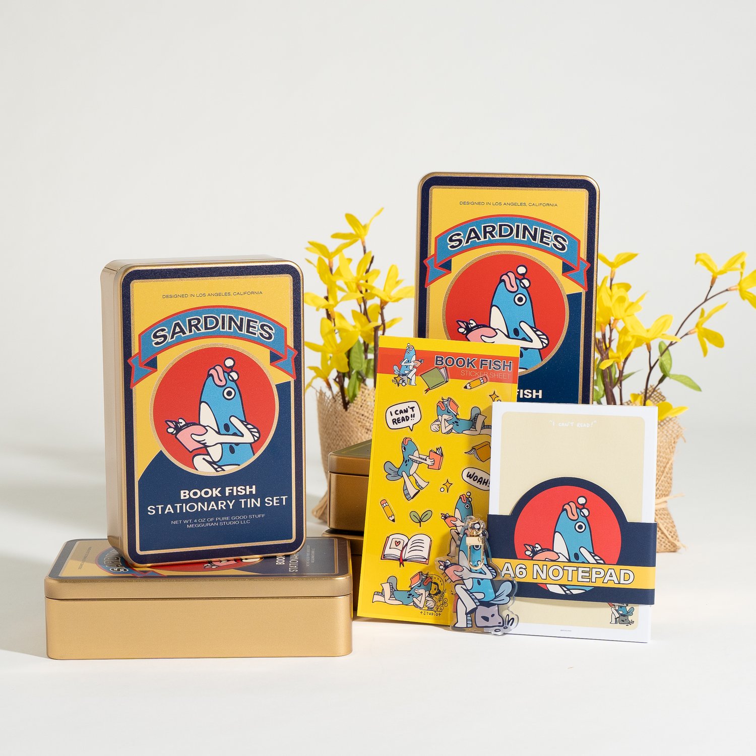 he image shows a collection of stationery items packaged in a retrostyle sardine tin.  the tin is yellow and gold and features a whimsical illustration of a fish. the set includes a notepad, stickers, and additional stationery items, all featuring a similar design aesthetic. yellow forsythia flowers are placed around the stationery, providing a bright and cheerful ambiance to the image. the entire scene is presented against a clean, white background.