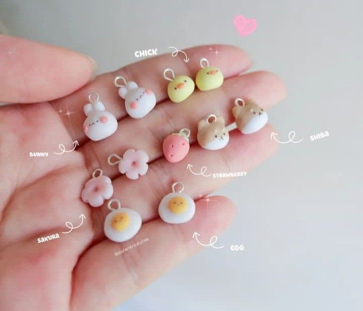 the image shows a hand holding a collection of small, handmade clay charms. the charms are in the shape of various cute and kawaiistyle objects, including a bunny, chick, shiba, strawberry, egg, and sakura (cherry blossom). they are small and colorful, and they appear to be jewelry or accessories components.