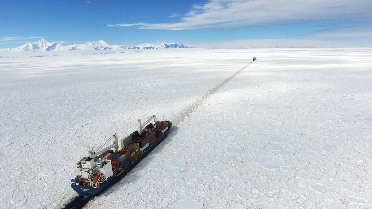 an aerial view of a container ship cutting through a frozen sea in antarctica. the ship is leaving a long trail in the ice, and the mountains in the distance are covered in snow. the sky is a bright blue, and the water is a deep blue.