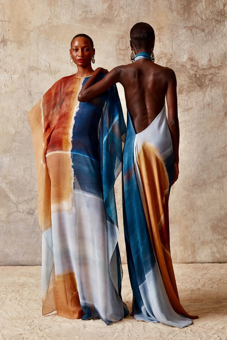 two women pose in the studio wearing similar patterned silk dresses. the woman in front is wearing a backless dress, and the woman behind her is wearing a dress with a high neckline. both dresses feature a geometric pattern with shades of blue, brown, and orange.