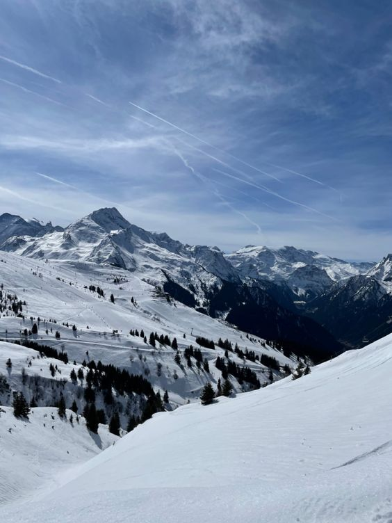 the image shows a snowy mountain landscape under a bright blue sky. slopes for skiing are visible on the mountains, dotted with trees. the perspective is from a high point, looking down a slope covered in untouched snow.