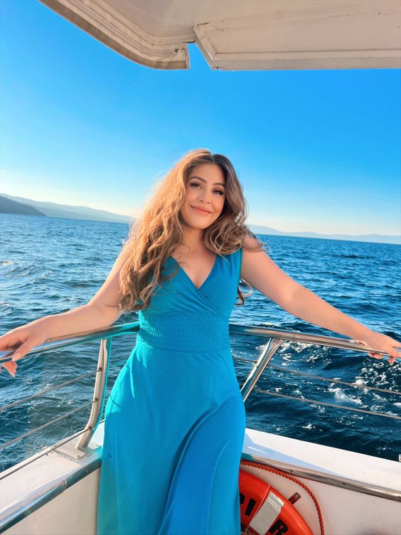 a woman in a long blue dress is standing on a boat and smiling at the camera. the water is blue, the sky is blue and the sun is shining. she is holding onto the railing of the boat with both hands. there is a life preserver behind her to her right.