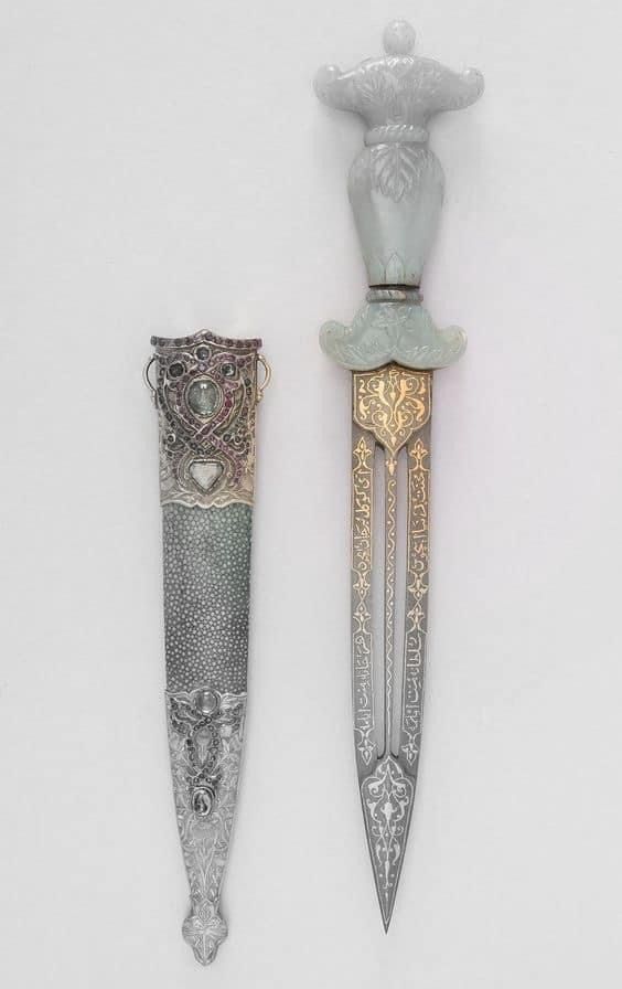 the image shows an ornate dagger with a sheath. the dagger's handle is made of a light green material, possibly jade, and is sculpted with decorative details. the blade is silver with gold inlays and ornate patterns. the sheath is primarily silver and appears to be inlaid with a stippled gray material, possibly shagreen. it also has decorative silver accents and embellishments.