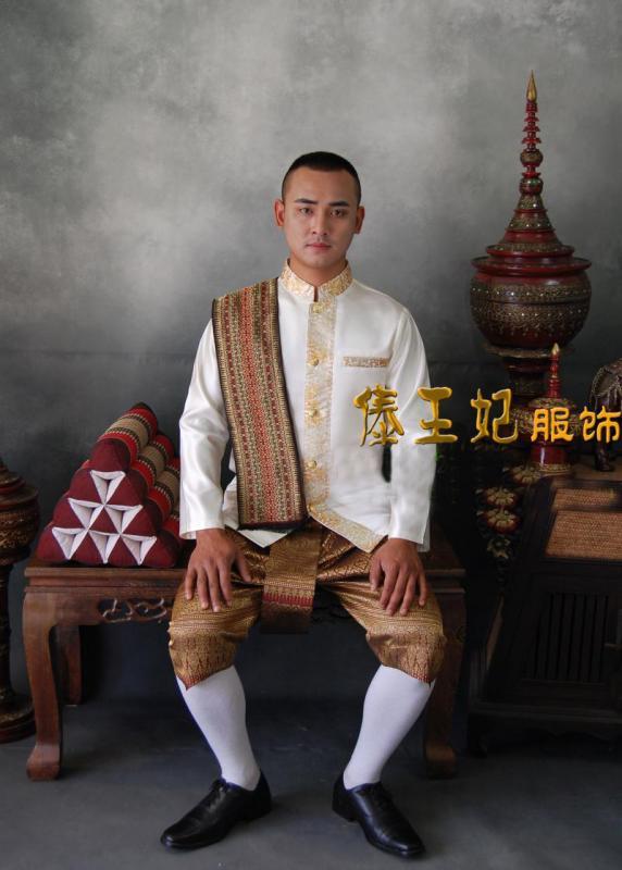 a man is sitting in traditional thai clothing. he is wearing a white, gold, and red silk ensemble. he has a gold, ornate scarf draped around his neck. his trousers are gold and red with a pattern. he has white socks and black shoes. the man is sitting on a chair with a red pillow and a brown wooden table. the background is a beige wall with a decorative vase and a small, decorative statue.