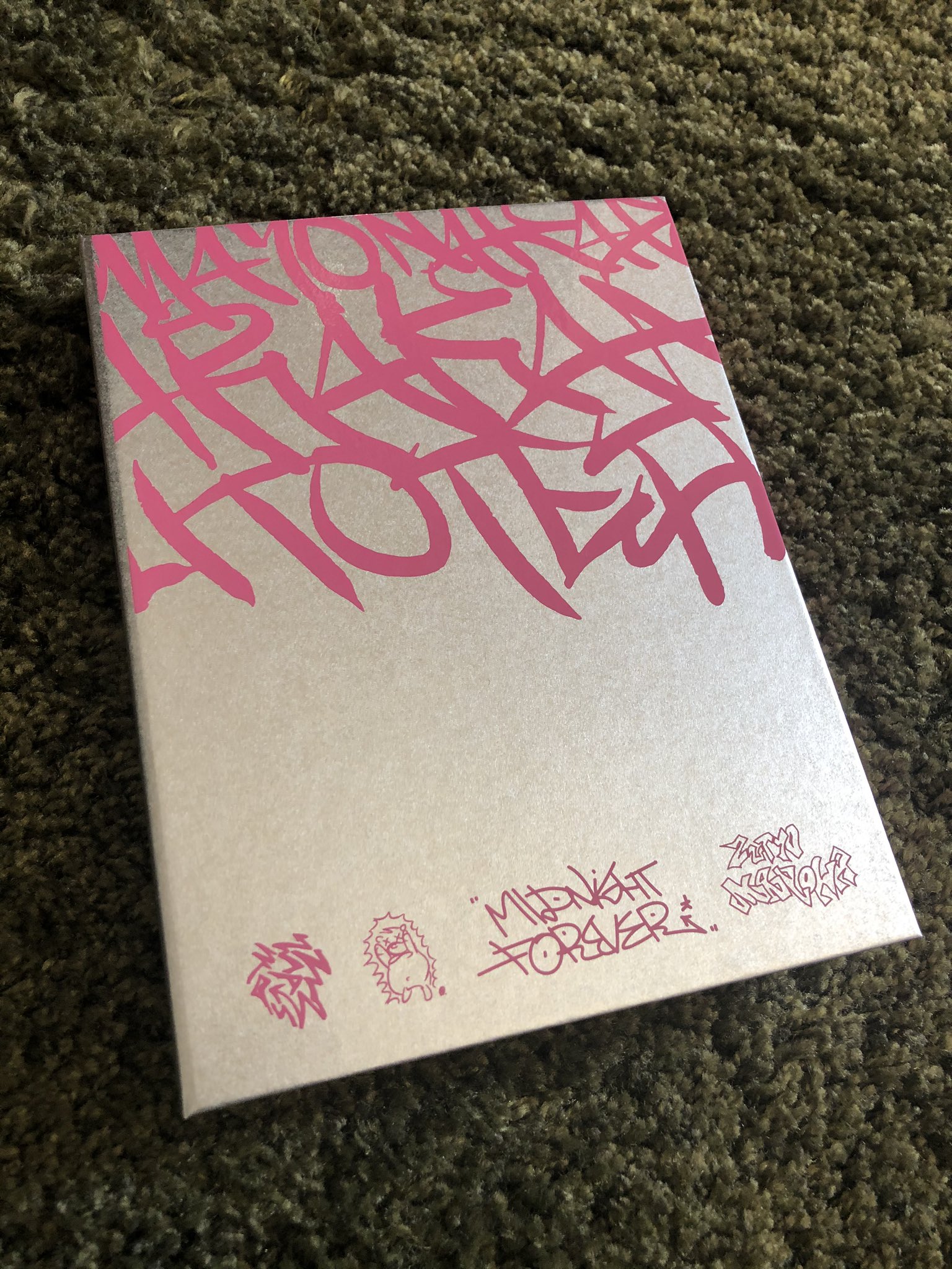 a silver rectangular box lays on a green textured surface. the box is slightly angled and features pink graffitistyle text that reads "midnight forever." in the bottom right corner. additional pink graphics and text are present on the box, with the most prominent text being large pink letters on a silver background that appear to spell "mason gucci beach hotel."  overall, the aesthetic appears to be inspired by street art and graphic design.
