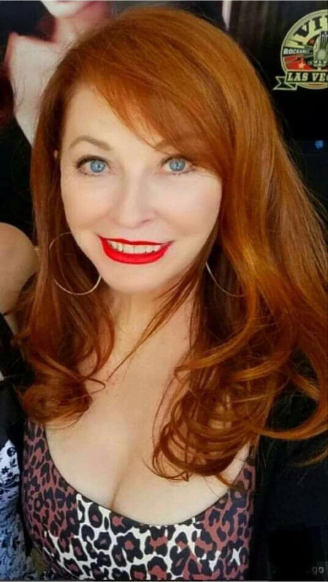 a closeup portrait of a smiling woman with long, red hair. she is wearing a leopard print top and has red lipstick on. she is looking at the camera with a happy expression.