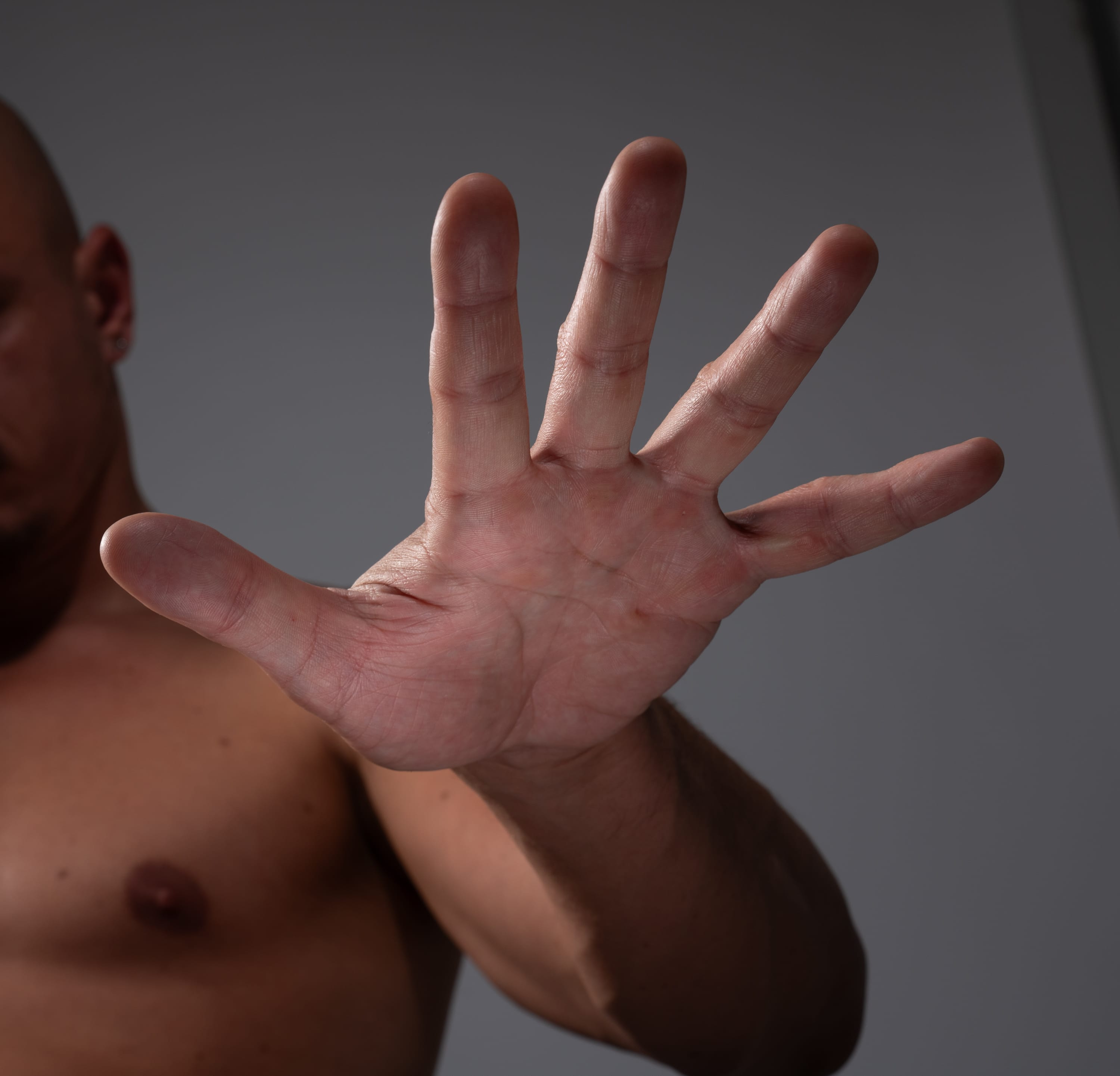 a man's hand is extended towards the viewer, palm out, in a gesture of "stop" or rejection. the man's bare torso is visible in the background. the background is a neutral gray color, and the lighting is soft and diffused. the image is a closeup shot, focusing on the hand and the gesture.