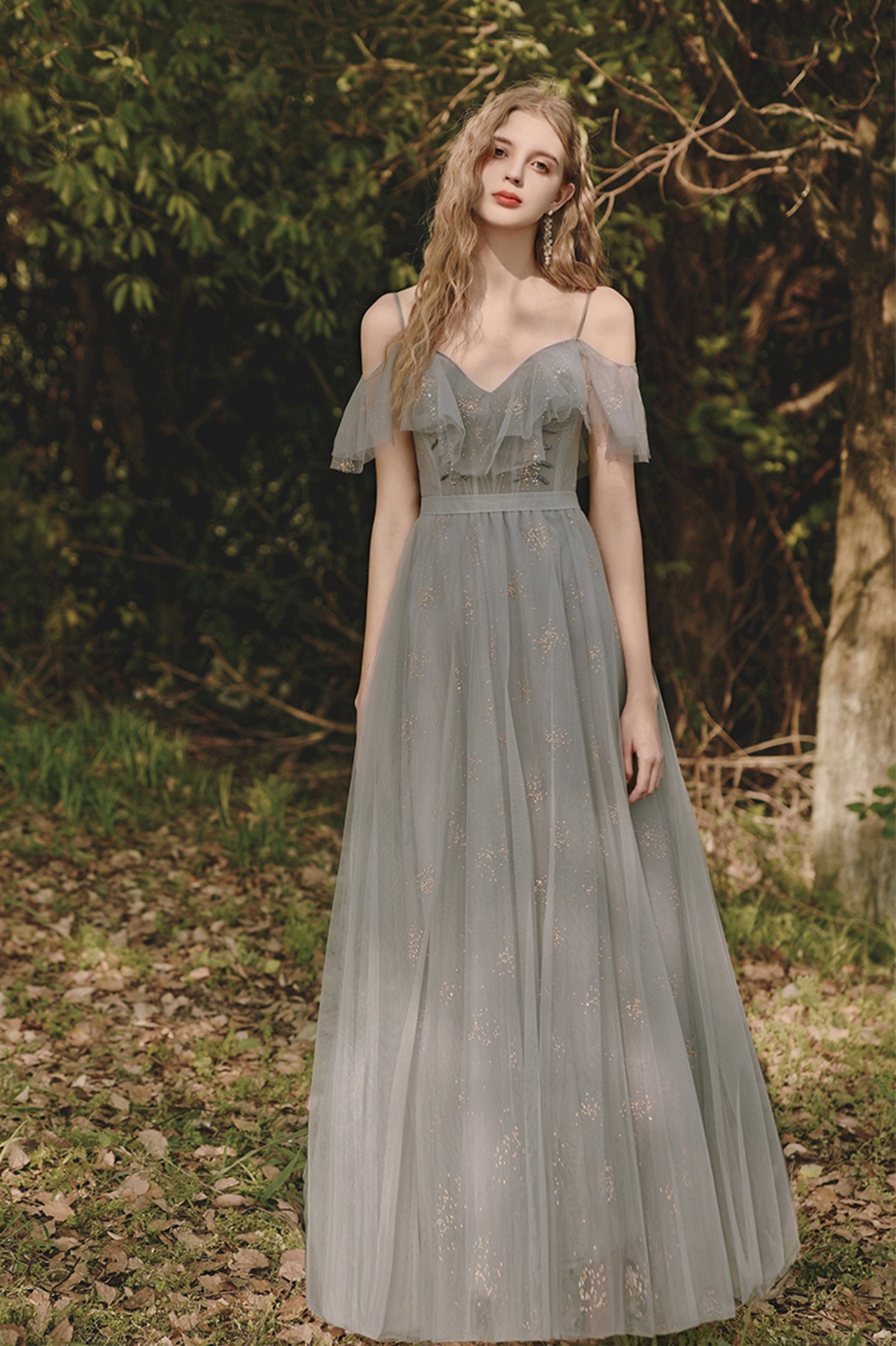 a young woman stands in a forest setting, wearing a long, gray tulle evening gown. the dress features delicate spaghetti straps, an offtheshoulder design with ruffles at the neckline, and a fitted bodice with a thin belt at the waist. the gown flows into a full, floorlength skirt adorned with subtle patterns. the woman has long, wavy blonde hair and is wearing dangling earrings. the background consists of trees and leaves, suggesting a natural, outdoor setting.
