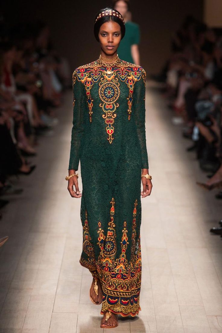 a darkskinned model walks the runway wearing a long, darkgreen lace dress adorned with intricate gold and multicolored embroidery. the dress has long sleeves and a high neckline. she is accessorized with a gold headband, gold bracelets and sandals.  the background features other models and spectators, typical of a fashion show setting. the overall aesthetic suggests a highfashion, possibly bohemian or ethnicinspired collection.