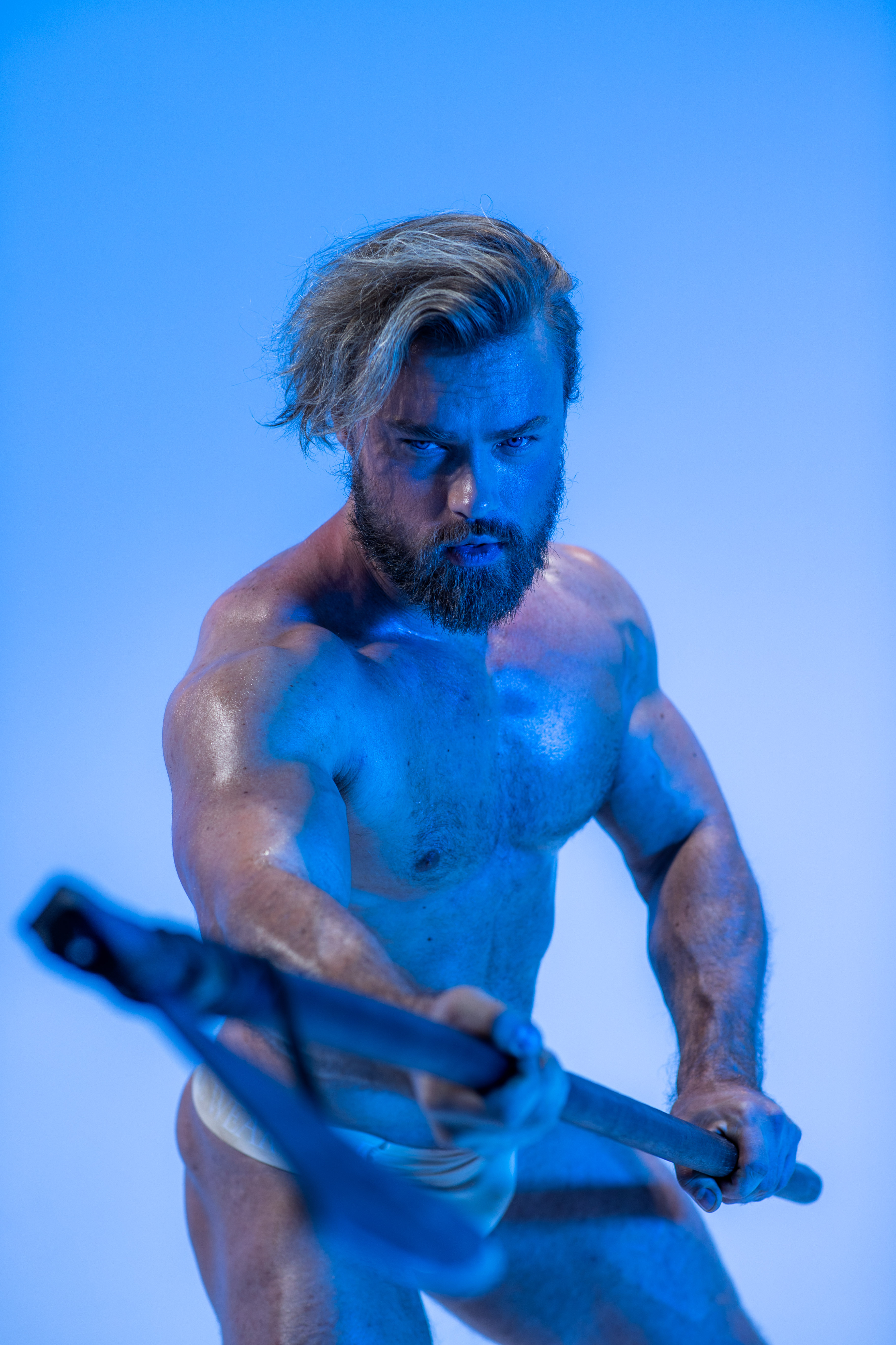 a muscular man with a beard, lit dramatically with blue light, stands holding a large, black axe. he appears to be midswing with a determined expression on his face. he is shirtless, showcasing his physique, and wearing minimal clothing. the background is a bright, almost electric blue, emphasizing the intensity of the scene.