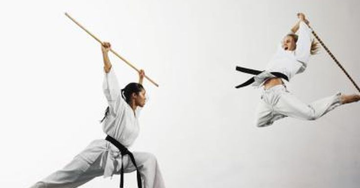 two women are practicing martial arts in a white studio. the woman on the left is holding a wooden staff and is in a low stance, ready to strike. the woman on the right is midair, performing a high kick with a black belt around her waist.