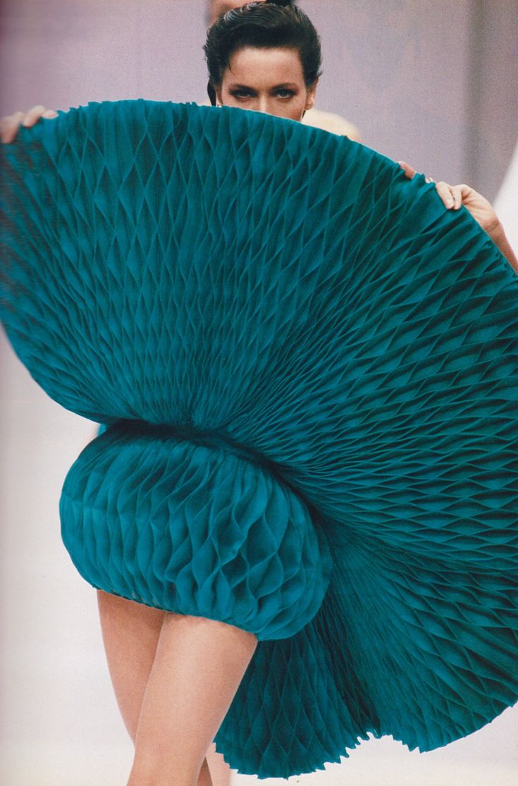 a woman models a teal dress with a honeycomb pleated design on a runway. the dress has a large circular pleated section that she is holding up in front of her face, with a spherical pleated section around her waist. the overall effect is dramatic and textured.