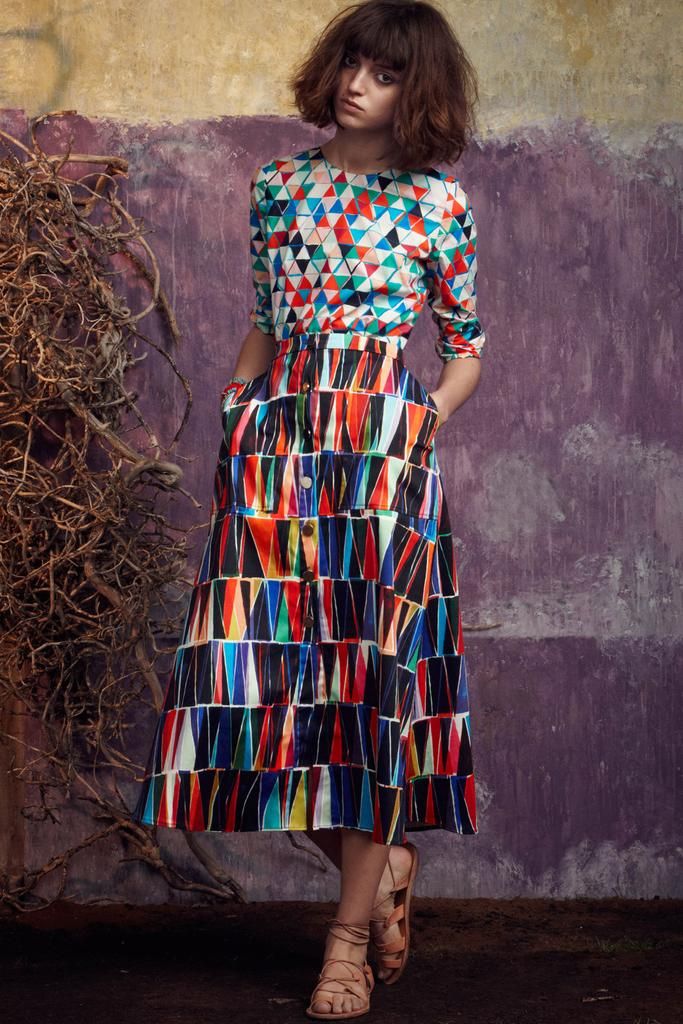 a fulllength shot of a young woman with short brunette hair, wearing a vibrant, geometricprinted dress. the dress consists of a fitted, threequartersleeved top and a flowing midi skirt, both featuring a pattern of multicolored triangles. she is standing against a textured purple wall with branches in the foreground. the woman is wearing brown strappy sandals and is posing with her hands in her pockets. the overall style is bohemian and retro.