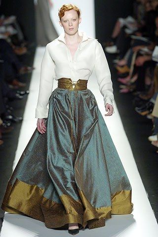 a redhead model walks down a runway, showcasing a teal and gold long gown. the dress consists of a flowing teal skirt with a gold band at the hem, paired with a crisp white blouse and a structured gold belt cinching the waist. the model's makeup is natural and her hair is styled in a loose updo. the background is blurred, showing the audience and other aspects of the fashion show setting. the overall image exudes high fashion and elegance.