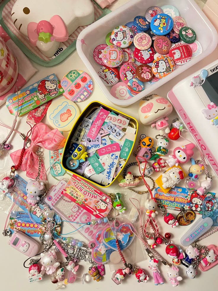 the image displays a collection of hello kitty merchandise and accessories, including buttons, charms, keychains, and other small trinkets. the items are arranged on a white surface, with some stored in containers such as a white tray and a yellow tin box. the predominant colors are pink, white, and yellow, with other accent colors like red, blue, and green also present. the items feature hello kitty characters and designs, and appear to be vintage or collectible items. the image suggests a collection of hello kitty memorabilia.