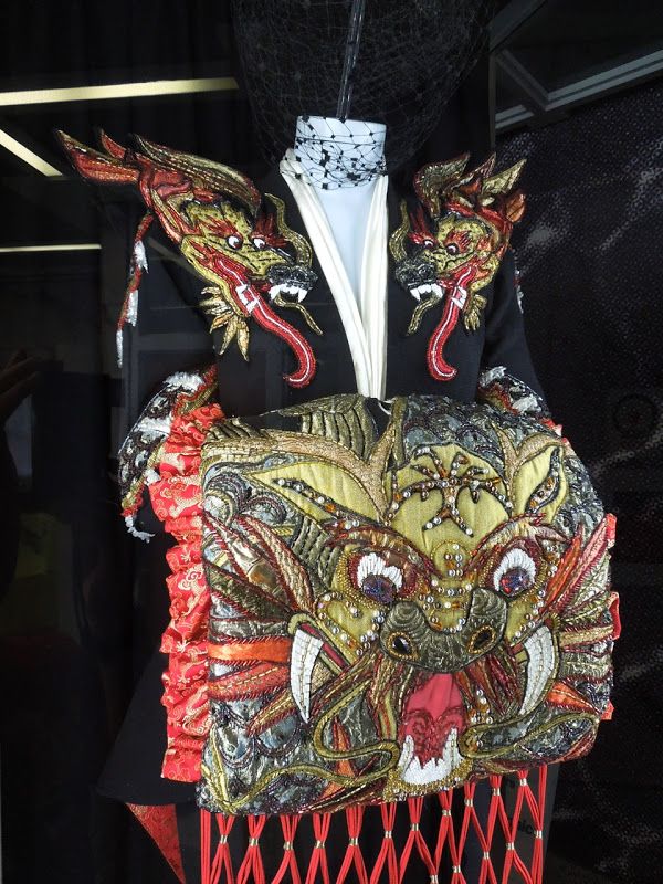 a theatrical costume is displayed, featuring elaborate dragon embroidery and ornamentation. the garment consists of a black jacket with large, red and gold dragon head shoulder pads, and a squareshaped front piece also adorned with a highly detailed dragon face. the front panel has tassels hanging from the bottom. red fabric ruffles accent the sleeves and sides. the costume is displayed on a mannequin within a display case.