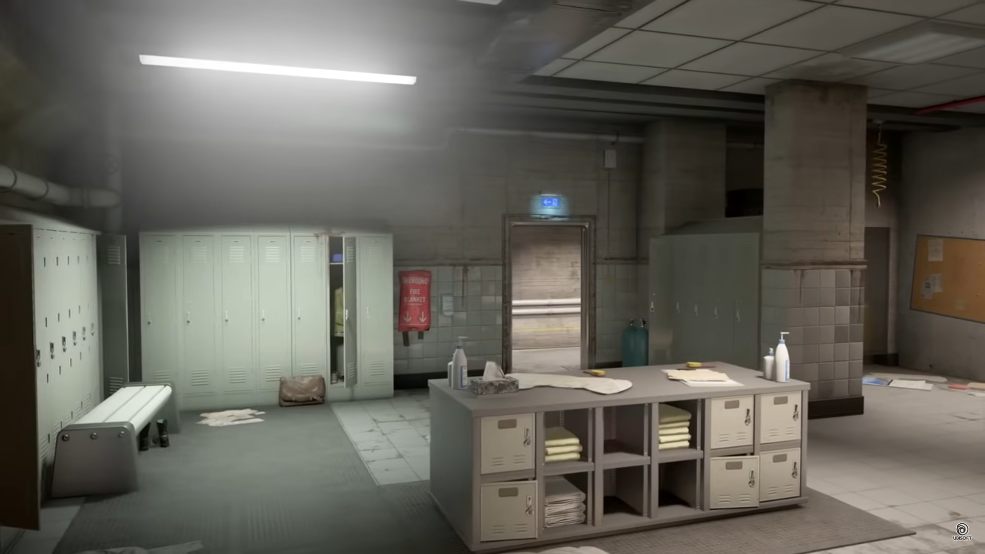 a locker room interior. there are multiple lockers on the left side of the image, a bench beneath them, a few towels and toiletries on a counter with more lockers below it, and a door leading to an outside hallway. the floor is tiled gray.