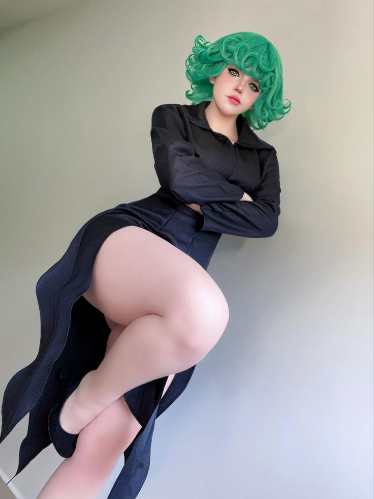 a woman dressed as tatsumaki from the anime onepunch man poses indoors against a plain wall. she wears a green wig, a black dress with a high slit, and black heels. her arms are crossed and she is looking at the camera. the image is taken from a low angle.